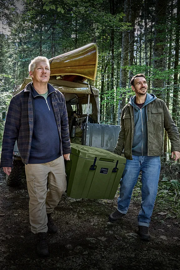 Zwei Männer tragen eine grüne Box über einen Waldweg; im Hintergrund ein Geländewagen mit Kanu.